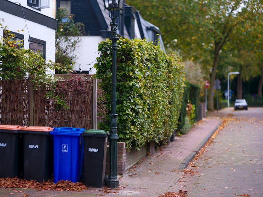 A suburban street scene featuring a paved sidewalk bordered by a well-maintained garden with dense, trimmed green hedges and trees with autumn-colored leaves. Along the sidewalk, there is a black orna