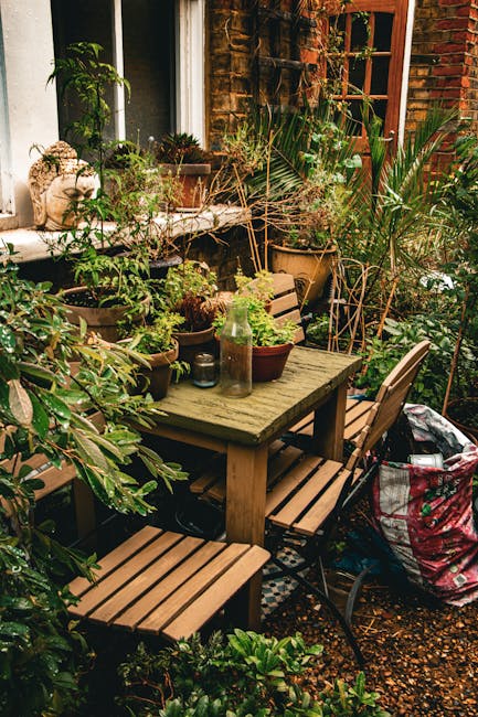 A cozy outdoor garden seating area featuring a rectangular wooden table surrounded by four matching wooden chairs, set against the backdrop of a brick wall and window. The table is decorated with smal